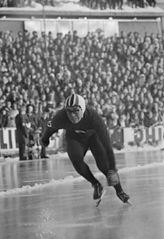A speed skater in a helmet and glasses is captured in motion on the ice, leaning forward with intensity. In the background, a large crowd watches the event, dressed in winter clothing.
