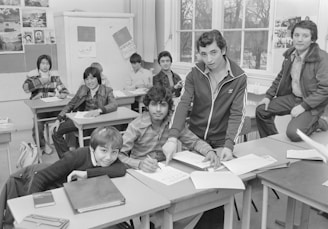 A classroom scene featuring a group of students sitting at desks. Some students are writing, while others are posing and looking towards the camera. The room is equipped with desks, papers, and a blackboard displaying various images and scripts. Windows are visible in the background, letting natural light in.