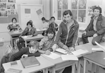 A classroom scene featuring a group of students sitting at desks. Some students are writing, while others are posing and looking towards the camera. The room is equipped with desks, papers, and a blackboard displaying various images and scripts. Windows are visible in the background, letting natural light in.