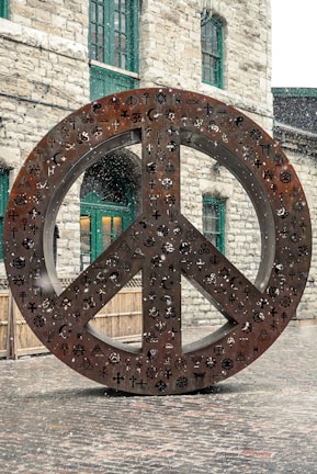 A large metal sculpture of a peace symbol stands outdoors, adorned with various religious and cultural symbols. Snow is gently falling around the sculpture, which is set against a backdrop of a stone building with green-framed windows.