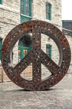 A large metal sculpture of a peace symbol stands outdoors, adorned with various religious and cultural symbols. Snow is gently falling around the sculpture, which is set against a backdrop of a stone building with green-framed windows.