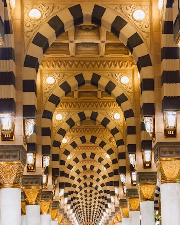A series of intricately designed arches with alternating black and beige stripes, adorned with ornate carvings and detailed patterns. The arches are illuminated with soft, warm lighting from hanging lanterns, creating a sense of depth and grandeur.