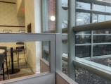 A modern indoor setting with a view through large glass windows. The interior features a dining area with round tables and dark chairs, separated by a partial glass partition. The exterior visible through the windows includes a building facade with reflective glass panels and a landscaped area.