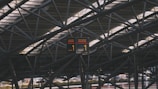 A complex network of steel beams creates an intricate pattern against the ceiling of a modern structure. A digital display sign hangs in the center, displaying the number 1 and indicating a terminal at Hamburg Airport.