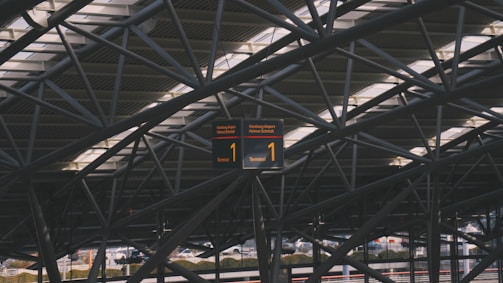 A complex network of steel beams creates an intricate pattern against the ceiling of a modern structure. A digital display sign hangs in the center, displaying the number 1 and indicating a terminal at Hamburg Airport.