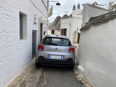 A compact car is parked in a narrow alleyway between whitewashed stone buildings. The buildings have distinctive conical roofs, likely indicating a traditional architectural style. The alley is very tight, just wide enough for the car. There is a street lamp hanging above, and one of the buildings has a wooden balcony.