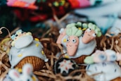 Cupcakes decorated with playful designs, featuring whimsical icing shapes resembling carrots and other colorful, cartoon-like figures. The cupcakes are arranged on shredded brown paper, creating a rustic presentation.