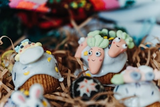 Cupcakes decorated with playful designs, featuring whimsical icing shapes resembling carrots and other colorful, cartoon-like figures. The cupcakes are arranged on shredded brown paper, creating a rustic presentation.