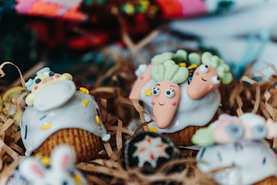Cupcakes decorated with playful designs, featuring whimsical icing shapes resembling carrots and other colorful, cartoon-like figures. The cupcakes are arranged on shredded brown paper, creating a rustic presentation.
