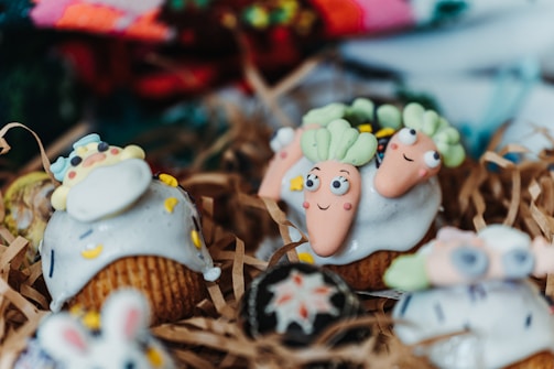 Cupcakes decorated with playful designs, featuring whimsical icing shapes resembling carrots and other colorful, cartoon-like figures. The cupcakes are arranged on shredded brown paper, creating a rustic presentation.