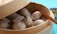 A bamboo steamer containing several steamed dumplings, partially open to reveal the contents. A pair of chopsticks rests on the edge of the steamer. In the background, there is a small bowl with sliced spring onions.