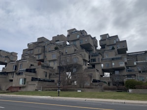 An architectural structure composed of stacked modular concrete units, creating a geometric and futuristic appearance. The building features numerous windows and terraces, surrounded by some sparse landscaping and trees.