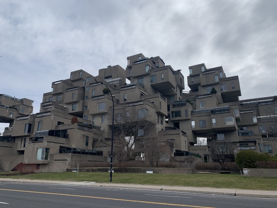 An architectural structure composed of stacked modular concrete units, creating a geometric and futuristic appearance. The building features numerous windows and terraces, surrounded by some sparse landscaping and trees.