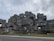 An architectural structure composed of stacked modular concrete units, creating a geometric and futuristic appearance. The building features numerous windows and terraces, surrounded by some sparse landscaping and trees.