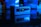 A small, rectangular computer device is set against a dark background. It is illuminated with a blue light and reflects on a glossy surface. The front panel features USB ports and an audio jack, while the blurred background shows indistinct shapes that imply a tech-themed setting.