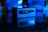 A small, rectangular computer device is set against a dark background. It is illuminated with a blue light and reflects on a glossy surface. The front panel features USB ports and an audio jack, while the blurred background shows indistinct shapes that imply a tech-themed setting.