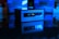 A small, rectangular computer device is set against a dark background. It is illuminated with a blue light and reflects on a glossy surface. The front panel features USB ports and an audio jack, while the blurred background shows indistinct shapes that imply a tech-themed setting.