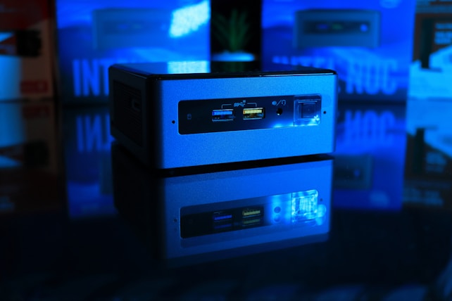 A small, rectangular computer device is set against a dark background. It is illuminated with a blue light and reflects on a glossy surface. The front panel features USB ports and an audio jack, while the blurred background shows indistinct shapes that imply a tech-themed setting.