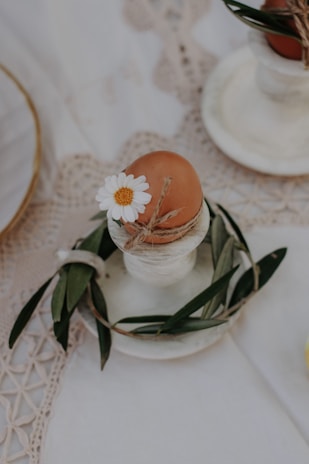 An egg with a brown shell is placed in a white egg cup. It is decorated with a small white daisy and wrapped with a twine string. Olive leaves are arranged around the base of the egg cup. The setup is on a lace-patterned cloth.