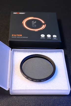A circular camera filter placed inside an open, white, protective case made of foam. The filter is labeled with specifications and brand details. Accompanying the filter is a black and orange product box with graphical elements and text indicating high-definition optical glass.