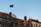 A European Union flag is waving atop a flagpole against a clear blue sky. The scene includes old-style buildings with ornate architectural details and chimneys. Leafless tree branches are visible in the foreground.
