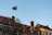 A European Union flag is waving atop a flagpole against a clear blue sky. The scene includes old-style buildings with ornate architectural details and chimneys. Leafless tree branches are visible in the foreground.
