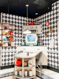 A vintage kitchen scene featuring patterned black and white wallpaper, a retro television, and various kitchen items. There is a small dining area with a corner table, chairs, and shelves displaying colorful jars, kitchenware, and a classic Coca-Cola bottle. Above the table, a pendant light hangs from the ceiling. The countertop holds some kitchen appliances and a second, smaller shelf is attached to the wall.