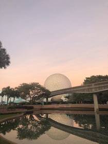 A large geodesic sphere is set against a soft, pastel sunset sky. Surrounding the sphere are lush trees and a monorail track. The scene is reflected in a calm body of water in the foreground.