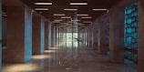 A long, symmetrical hallway with marble floors, flanked by tall stone columns. The ceiling features a grid-like pattern with integrated lighting, creating a modern and sleek look. On the right, a series of blue illuminated glass blocks adds a cool tone to the space, while natural light pours in from the far end.