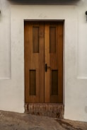 A sturdy wooden door with vertical rectangular glass panels set within a white wall. The door features a black handle on the right side and rests above a section of exposed brick. The surrounding wall has slight dirt marks, providing a rustic look.