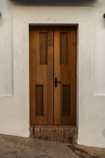 A sturdy wooden door with vertical rectangular glass panels set within a white wall. The door features a black handle on the right side and rests above a section of exposed brick. The surrounding wall has slight dirt marks, providing a rustic look.