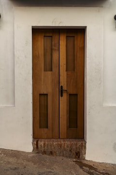 A sturdy wooden door with vertical rectangular glass panels set within a white wall. The door features a black handle on the right side and rests above a section of exposed brick. The surrounding wall has slight dirt marks, providing a rustic look.
