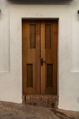 A sturdy wooden door with vertical rectangular glass panels set within a white wall. The door features a black handle on the right side and rests above a section of exposed brick. The surrounding wall has slight dirt marks, providing a rustic look.