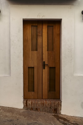 A sturdy wooden door with vertical rectangular glass panels set within a white wall. The door features a black handle on the right side and rests above a section of exposed brick. The surrounding wall has slight dirt marks, providing a rustic look.