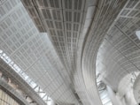 Curved and linear metal structures form an intricate roof design. The architecture appears modern with a focus on geometric patterns and clean lines. The ceiling panels reflect light, adding a sense of brightness and spaciousness to the area.