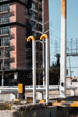 A street scene featuring tall, vertical metal pipes with yellow curves at the top, set against the backdrop of a modern brick and glass building. The foreground includes concrete and metal structures, suggesting an urban setting. Power lines and an electrical grid frame the sky, adding to the urban landscape.