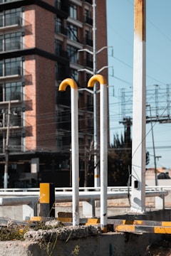 A street scene featuring tall, vertical metal pipes with yellow curves at the top, set against the backdrop of a modern brick and glass building. The foreground includes concrete and metal structures, suggesting an urban setting. Power lines and an electrical grid frame the sky, adding to the urban landscape.