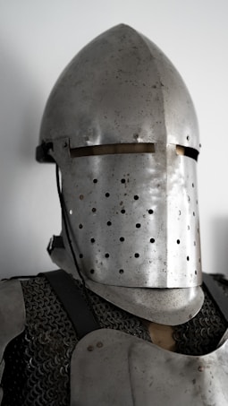 A close-up of a medieval knight's helmet and chainmail armor. The helmet is metallic and has a conical shape with small round perforations across its surface. The chainmail is visible beneath the helmet, and the entire armor has a slightly worn, antique look.