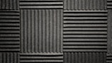 A pattern of black acoustic foam panels arranged in a grid. Each panel features alternating horizontal and vertical grooves, creating a textured appearance that absorbs sound. The lighting emphasizes the texture and depth of the grooves.
