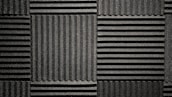 A pattern of black acoustic foam panels arranged in a grid. Each panel features alternating horizontal and vertical grooves, creating a textured appearance that absorbs sound. The lighting emphasizes the texture and depth of the grooves.