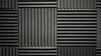 A pattern of black acoustic foam panels arranged in a grid. Each panel features alternating horizontal and vertical grooves, creating a textured appearance that absorbs sound. The lighting emphasizes the texture and depth of the grooves.