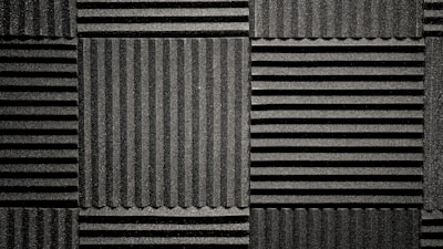 A pattern of black acoustic foam panels arranged in a grid. Each panel features alternating horizontal and vertical grooves, creating a textured appearance that absorbs sound. The lighting emphasizes the texture and depth of the grooves.