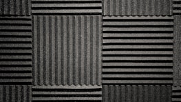 A pattern of black acoustic foam panels arranged in a grid. Each panel features alternating horizontal and vertical grooves, creating a textured appearance that absorbs sound. The lighting emphasizes the texture and depth of the grooves.