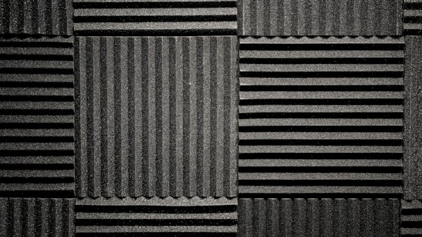 A pattern of black acoustic foam panels arranged in a grid. Each panel features alternating horizontal and vertical grooves, creating a textured appearance that absorbs sound. The lighting emphasizes the texture and depth of the grooves.