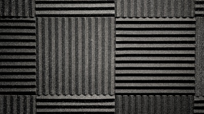 A pattern of black acoustic foam panels arranged in a grid. Each panel features alternating horizontal and vertical grooves, creating a textured appearance that absorbs sound. The lighting emphasizes the texture and depth of the grooves.