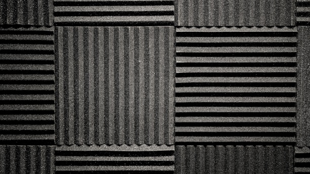 A pattern of black acoustic foam panels arranged in a grid. Each panel features alternating horizontal and vertical grooves, creating a textured appearance that absorbs sound. The lighting emphasizes the texture and depth of the grooves.