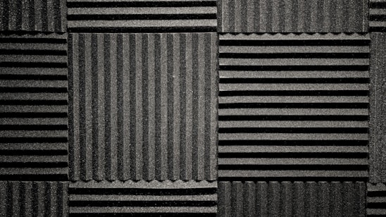 A pattern of black acoustic foam panels arranged in a grid. Each panel features alternating horizontal and vertical grooves, creating a textured appearance that absorbs sound. The lighting emphasizes the texture and depth of the grooves.