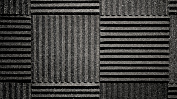 A pattern of black acoustic foam panels arranged in a grid. Each panel features alternating horizontal and vertical grooves, creating a textured appearance that absorbs sound. The lighting emphasizes the texture and depth of the grooves.