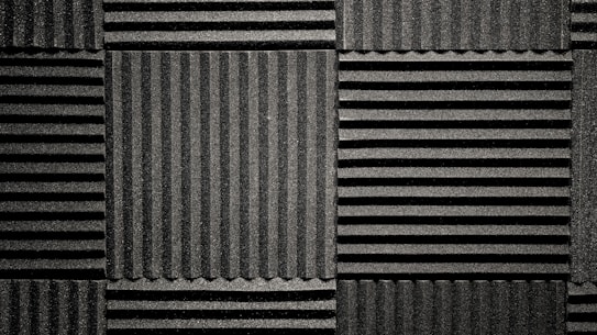 A pattern of black acoustic foam panels arranged in a grid. Each panel features alternating horizontal and vertical grooves, creating a textured appearance that absorbs sound. The lighting emphasizes the texture and depth of the grooves.