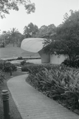 A curving pathway leads through a landscaped garden area filled with dense foliage and plants. The path approaches a modern, uniquely shaped building with a ribbed roof. Tall trees and a well-manicured lawn are visible in the background, evoking a serene and natural environment.